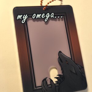 May include: A rectangular tag with a brown border and the words "my omega..." in white script. A black wolf illustration with orange eyes and claws is on the right side. A small orange beaded chain is attached at the top.