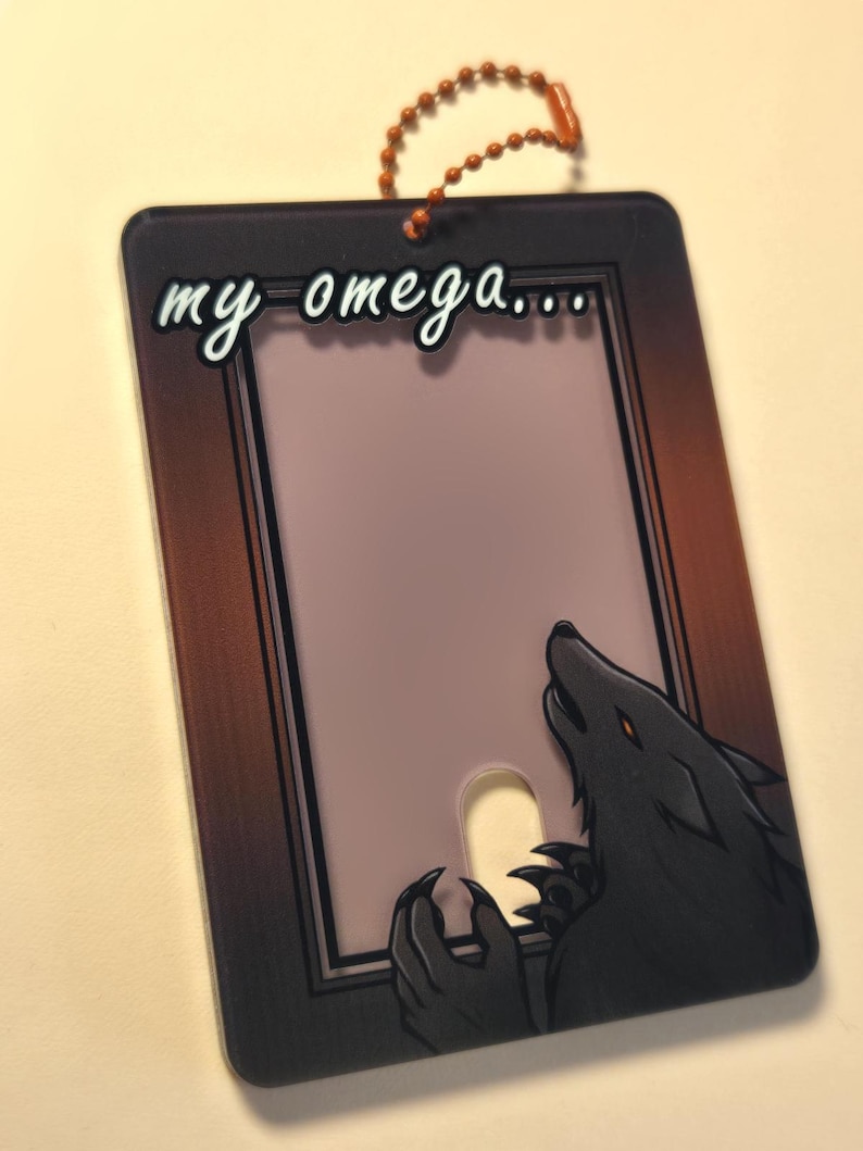 May include: A rectangular tag with a brown border and the words "my omega..." in white script. A black wolf illustration with orange eyes and claws is on the right side. A small orange beaded chain is attached at the top.