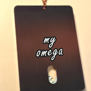 May include: A rectangular tag with a gradient brown background and the words "my omega" in white script. The tag has a small cutout and is attached to a beaded chain.
