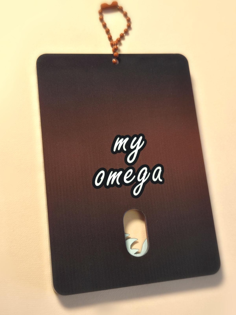 May include: A rectangular tag with a gradient brown background and the words "my omega" in white script. The tag has a small cutout and is attached to a beaded chain.
