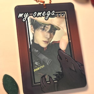 May include: A rectangular keychain with a photo of a person and a wolf illustration. The keychain has the text "my-omega..." in white cursive. The photo is framed in a dark brown border, and the keychain has a small orange beaded chain.