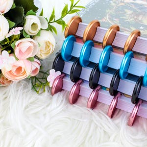 Może przedstawiać: Stos notesów z kolorowymi metalowymi pierścieniami do wiązania. Pierścienie są w odcieniach miedzi, niebieskiego, czarnego i różowego.