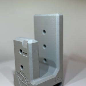 May include: A light grey, 3D-printed object with a geometric design and the text "AR-15". The object has multiple holes and a diamond-shaped pattern. It is designed to hold a specific item.