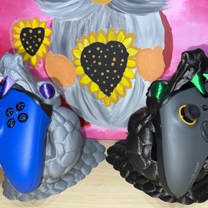 May include: Two 3D printed game controller holders, one blue and one black, shaped like dragons with open mouths. The holders are designed to hold Xbox controllers.