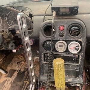 Puede incluir: Un interior de coche con una palanca de cambios alta de metal plateado con recortes ovalados, un pomo amarillo con patrón de grietas, una radio Uniden negra, una unidad Pioneer y tres indicadores analógicos circulares en la consola central.