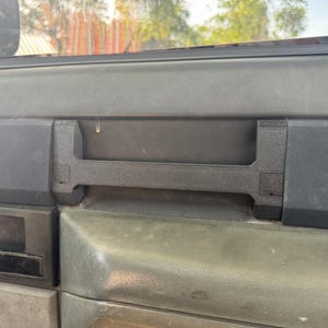 May include: A close-up of a black plastic dashboard panel with a handle. The panel has a textured surface and is likely from a car.