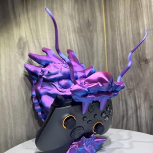 May include: A purple and blue iridescent dragon head controller stand with a black Xbox controller. The dragon has a textured surface and is holding the controller in its mouth.