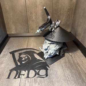 May include: A black airbrush rests on a black stand shaped like a Japanese demon mask. A gray demon head is below the airbrush. A black metal cutout of a grim reaper with the text "NFDG" is on the left.