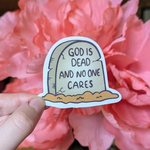 May include: A sticker in the shape of a tombstone with the words "GOD IS DEAD AND NO ONE CARES" in black lettering. The tombstone is light beige with a dark blue outline, set against a pink floral background.
