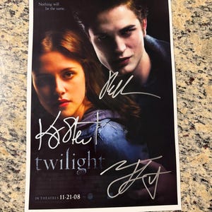以下が含まれることがあります： 暗い髪の二人の人物のポートレート、「Nothing will be the same」、「twilight」、「IN THEATRES 11-21-08」というテキストが記載された長方形のトワイライト映画ポスターで、プリントの表面には銀色のインクで書かれた3つの明確なサインがあります。
