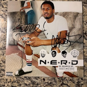 Puede incluir: Una portada de álbum de música cuadrada que muestra a una persona con una camiseta blanca sosteniendo un mando de juego mientras está sentada en un sofá. La portada incluye firmas en tinta negra, el texto N*E*R*D IN SEARCH OF... y una etiqueta Parental Advisory.