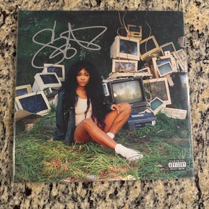 May include: A square music album cover featuring a person with long dark hair sitting in tall green grass surrounded by a pile of vintage beige computer monitors. A silver signature is written on the top left and a Parental Advisory label is on the bottom right.