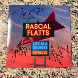 May include: A square album cover featuring a sunset sky background with a red neon sign reading RASCAL FLATTS and a blue and orange sign reading Duets LIFE IS A HIGHWAY REFUELED. The cover includes three distinct black ink signatures across the surface.