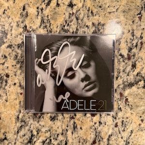 Puede incluir: Una caja de CD en blanco y negro que presenta un retrato de Adele con un autógrafo blanco escrito a mano en la portada. El texto ADELE 21 está impreso en la esquina inferior derecha. La caja es de plástico transparente y contiene el álbum.