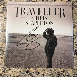 Könnte beinhalten: Ein quadratisches Vinyl-Schallplattencover für das Album Traveller von Chris Stapleton. Das Cover zeigt ein Schwarz-Weiß-Foto einer Person mit Cowboyhut und karierter Jacke in einer Wüstenlandschaft. Auf der Vorderseite befindet sich eine schwarze Tintensignatur.
