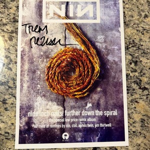 May include: A signed Nine Inch Nails "Further Down the Spiral" album cover. The image features the band's logo, a spiral design, and the text "nine inch nails: further down the spiral". The album cover is signed by a band member.