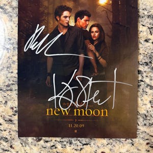 Puede incluir: Un póster de película rectangular de The Twilight Saga: New Moon con tres actores en un entorno forestal con una luna brillante. El póster incluye el texto "the twilight saga new moon" y "11.20.09" con dos firmas distintas en tinta blanca.