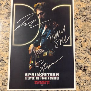 May include: A rectangular movie poster for Springsteen Deliver Me From Nowhere featuring a person playing an electric guitar, three white ink signatures, the Dolby Cinema logo, and the text Only in Theaters October 24 on a dark background.