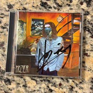 May include: A Hozier CD case featuring a surreal illustration of a person in a light blue button-down shirt with a landscape replacing their face. The artwork includes a clock, a staircase, and a desk. A bold black autograph is across the front.
