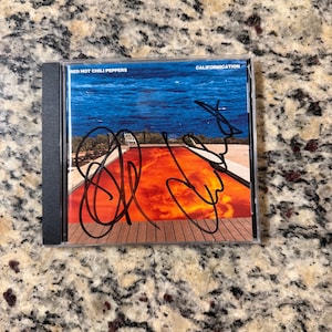 May include: A Red Hot Chili Peppers Californication CD case featuring a swimming pool with orange water and a blue sky, with black ink signatures across the front cover. The text Red Hot Chili Peppers and Californication is printed on the cover.