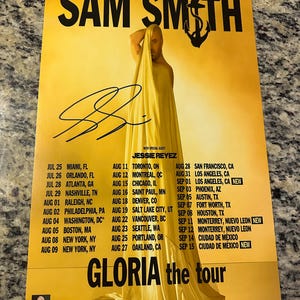 May include: A yellow concert poster with a black anchor and the text "SAM SMITH" in large black letters. The poster advertises the "GLORIA the tour" with dates and cities for the tour. The poster features a photo of Sam Smith wearing a yellow sheet and a signature.