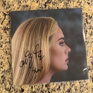 Puede incluir: Una portada de álbum de música cuadrada con un retrato de perfil de una persona rubia mirando hacia la derecha. La imagen incluye un autógrafo en tinta negra sobre el cabello. El fondo es de tono gris oscuro y el objeto está en plástico.