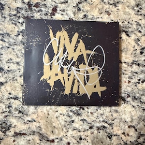 Puede incluir: Una caja de CD cuadrada de color negro con un diseño de salpicaduras de pintura dorada y el texto VIVA LA VIDA impreso en una fuente dorada estilizada. Una firma manuscrita blanca se superpone en el centro de la portada, contrastando con el fondo.