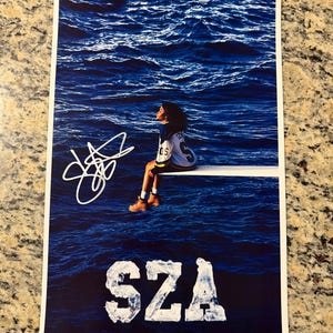 May include: A rectangular poster featuring a person sitting on a diving board over deep blue ocean water. The person wears a white and blue jersey with tan boots. A white signature is on the left, and the white text SZA is at the bottom.