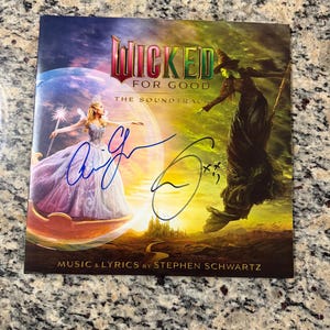 May include: A Wicked For Good soundtrack CD cover featuring a blonde figure in a light gown and a green-skinned figure in a dark dress, with two blue ink signatures across the front. The text reads Wicked For Good The Soundtrack Music & Lyrics by Stephen Schwartz.