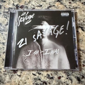Op de afbeelding: Een zwart-wit cd-hoesje voor het album I AM > I WAS van 21 Savage. De hoes toont een portret met de ogen bedekt door witte penseelstreken, de artiestennaam 21 Savage, de albumtitel en een Parental Advisory Explicit Content-label.