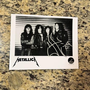 May include: Black and white photograph of the band Metallica. The band members are standing in front of a metal shutter door. The Metallica logo is at the bottom left corner of the photo.