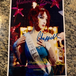 May include: A rectangular poster featuring a person with red hair wearing a blue dress and a white sash labelled CHAPPEL. The text THE RISE AND FALL OF A MIDWEST PRINCESS appears at the top, with a blue ink signature across the centre of the image.