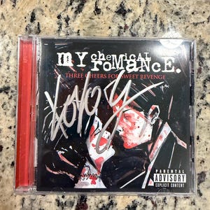 May include: A My Chemical Romance Three Cheers for Sweet Revenge CD in a clear plastic jewel case. The cover features a black and white illustration with red accents, a white signature across the front, and a Parental Advisory label.