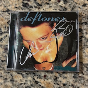 May include: A Deftones Around the Fur CD case featuring a high-angle photograph of a person in a black bra and white underwear with a flower tattoo on their stomach. The cover includes white ink signatures across the image and the text Deftones around the fur.