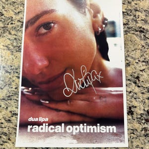 May include: A rectangular promotional print featuring a portrait of Dua Lipa with a silver autograph across the center. The text "dua lipa" and "radical optimism" appear in white at the bottom, with a small Warner Records logo below the text.
