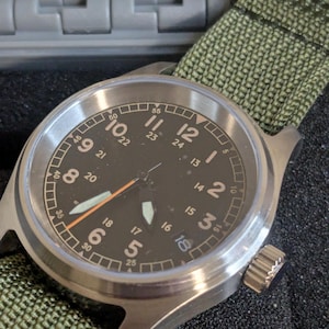 Op de afbeelding: Een roestvrijstalen polshorloge met een zwarte wijzerplaat en witte cijfers. Het horloge heeft een olijfgroene nylon band. De wijzers zijn wit en oranje. De wijzerplaat geeft zowel 12- als 24-uurs tijd weer. Militaire stijl.