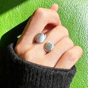 Puede incluir: Dos anillos de plata con un diseño liso y circular se muestran en una mano. Los anillos tienen un acabado pulido y están colocados sobre un fondo verde. La persona lleva un jersey negro.