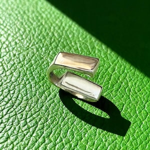 Könnte beinhalten: Ein silberfarbener Ring mit modernem Design. Der Ring hat zwei rechteckige Stäbe, die durch ein gebogenes Band verbunden sind. Er liegt auf einer strukturierten grünen Oberfläche, mit Schatten, die Tiefe verleihen.
