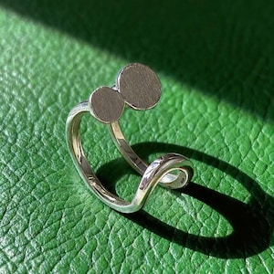 Pode incluir: Um anel de prata com um design único. O anel apresenta dois pequenos elementos circulares e planos e uma banda curva. O anel é mostrado numa superfície verde texturizada, com uma sombra.