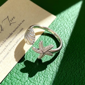 May include: A silver ring with a seashell and starfish design. The ring's band curves around the finger, with a seashell and a starfish as the main features. The starfish is decorated with tiny, sparkling details. The ring is on a green surface.