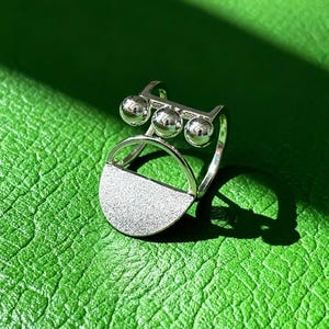 May include: A silver ring with a distinctive design. It has three small, spherical beads joined by a bar, and a half-circle shape with a textured surface. The ring is set against a green background.