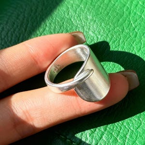 Könnte beinhalten: Ein silberner Ring mit einem einzigartigen Design, mit einem gebogenen Band und einer flachen, rechteckigen Fläche. Der Ring wird vor einem grünen Hintergrund gehalten, was seine polierte Metalloberfläche zeigt. Der Ring ist ein Schmuckstück.
