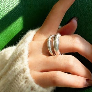 May include: A silver ring with a triple-band design. The ring is smooth and shiny, reflecting light. The ring is worn on a finger, with a cream-colored sweater in the background.