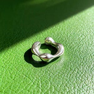 May include: A silver-colored ear cuff with an organic, flowing design. The cuff has a smooth, polished finish and a unique, sculptural shape. The ear cuff is shown on a green textured surface.