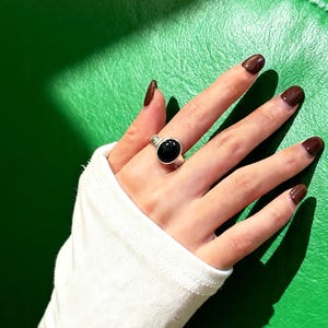 May include: A silver ring featuring a large, oval black gemstone. The ring is worn on a hand with brown nail varnish, set against a green backdrop. The ring's design is simple and elegant, suitable for everyday wear.