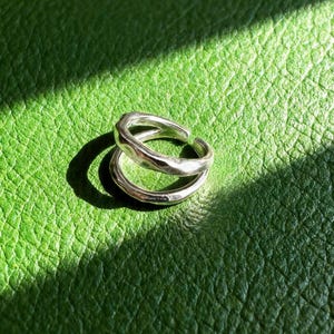 May include: A silver ring with a textured, organic design. The ring features two intersecting bands, creating a layered effect. It sits on a green, textured surface, with shadows adding depth to the image.