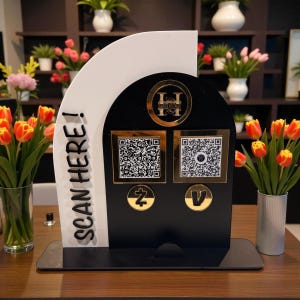 Op de afbeelding: Een zwart-witte QR-codestandaard met gouden accenten. De standaard heeft de tekst "SCAN HERE!" en twee QR-codes. Goudkleurige logo's en symbolen zijn ook aanwezig. De standaard staat op een houten oppervlak, met bloemen op de achtergrond.