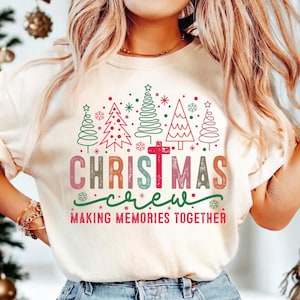 May include: Cream-colored Christmas crewneck t-shirt with a festive design. The graphic features Christmas trees, snowflakes, and the words "CHRISTMAS crew MAKING MEMORIES TOGETHER" in various colors. A red cross is in the center of the design.