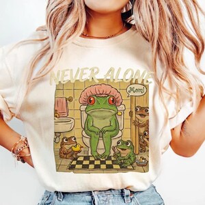 A woman wearing a white t-shirt with a graphic design depicting a green frog sitting on a toilet in a bathroom, surrounded by other frogs.