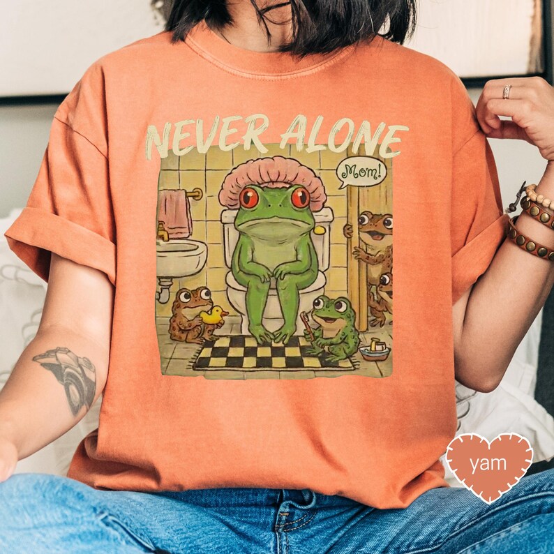 a person wearing an orange t-shirt with a cartoon image of a frog sitting on a toilet surrounded by other cartoon frogs.